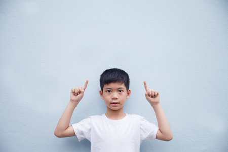 An Asian boy wearing a white shirt is pointing to the side in a blank space for text, against a blue...