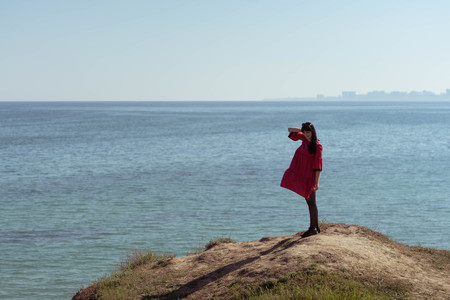 Beautiful brunette woman in red dress posing against the backdrop of the sea on a sunny spring day.の写真素材