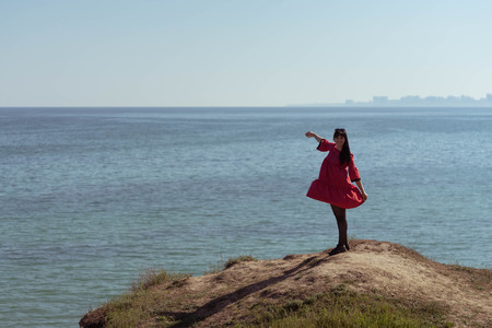 Beautiful brunette woman in red dress posing against the backdrop of the sea on a sunny spring day.の写真素材