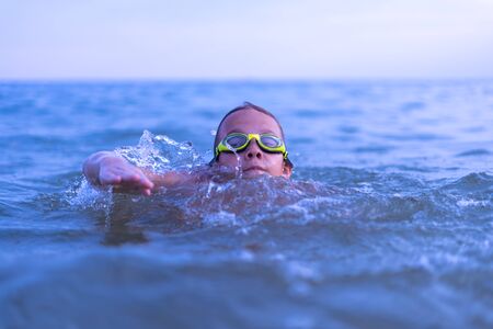 A 10-year-old boy swims in the sea at dawn with glasses for swimmingの写真素材