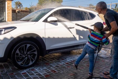 A middle-aged man teaches children of boys 4 and 10 years old to wash a car in the yard of his house on a summer sunny day. 2019.09.22. Odessa. Ukraineの写真素材