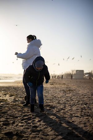 A 4 year old boy is playing and having fun on the seashore on a sunny winter day, while his mother, a brunette woman, photographs nature and the seaの写真素材
