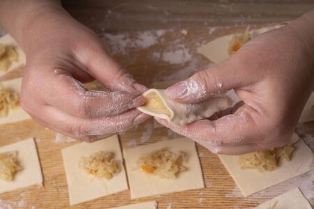 A woman sculpts dumplings and ravioli from squares of dough and cabbage. Plywood cutting board, wooden flour sieve and wooden rolling pin - tools for making doughの写真素材
