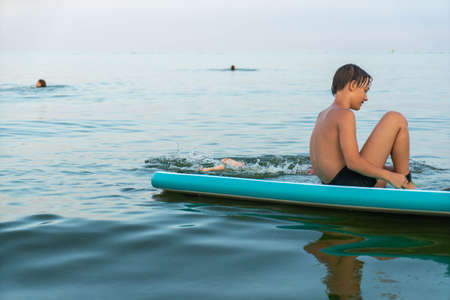 A boy of 11 years old swims on a SUP board in the sea after sunsetの写真素材