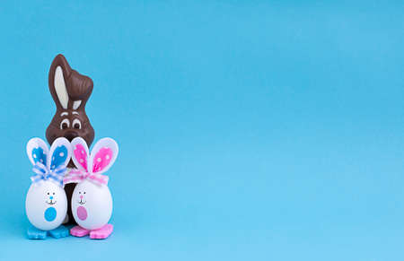 Chocolate bunny and two Easter eggs with painted eyes and rabbit ears and paws on a blue background...