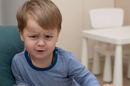 A small Caucasian handsome boy emotionally frowns his eyebrows in a home interior in a striped sweater. Close-upの写真素材