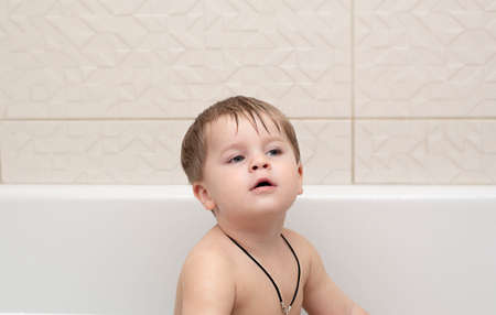 A small handsome caucasian boy is sitting in the bathroom and looking at the camera.