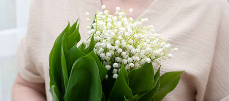 A woman in a beige sweater holds a bouquet of white lilies of the valley with bright green leaves in her hand.の写真素材