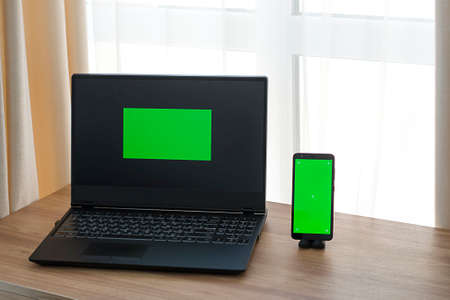 Chromakey. Laptop and phone with green screen on wooden table in home interior.