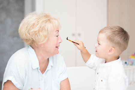 people. family concept. A small handsome Caucasian boy cheerfully paints his grandmother's face with paints. home interior. close-up. soft focus.の写真素材