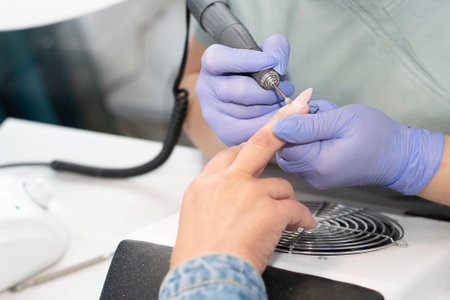 Manicurist in lilac latex gloves removes yellow gel polish from long nails to a client in a beauty...