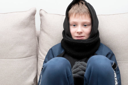 People. Energy crisis concept. A teenage boy sits at home on the couch in gloves and a hood, freezes...