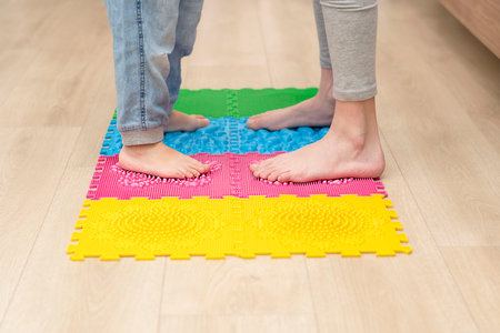 health concept. Two boys stand barefoot on a colorful orthopedic massage mat in a home interior. Massage and improve circulation of the feet. Close-up of children's legs.の写真素材