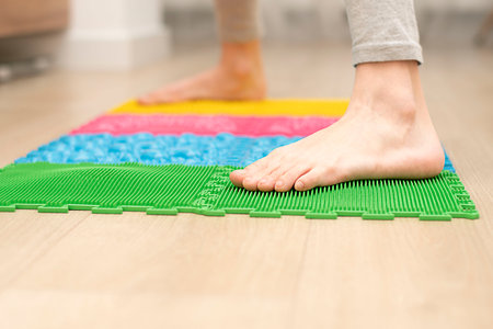 people. concept. healthy lifestyle. a teenager boy stands on a multi-colored orthopedic massage mat with bare feet in a home interior. It exercises the feet to improve blood circulation. Treatment of flat feet. close-up. soft focus.の写真素材