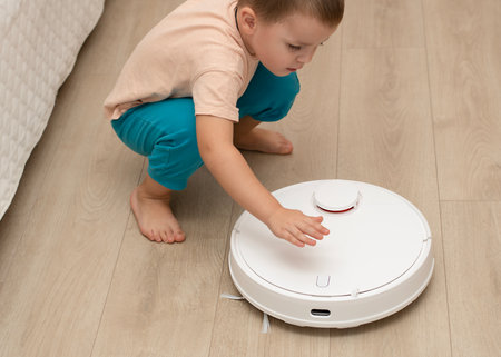 Cleaning concept. A little cheerful boy in home clothes, 4 years old, throws torn paper into small pieces on the floor for cleaning and suction by a white robot vacuum cleaner.の写真素材