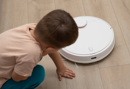 Cleaning concept. A little cheerful boy in home clothes, 4 years old, throws torn paper into small pieces on the floor for cleaning and suction by a white robot vacuum cleaner.の写真素材
