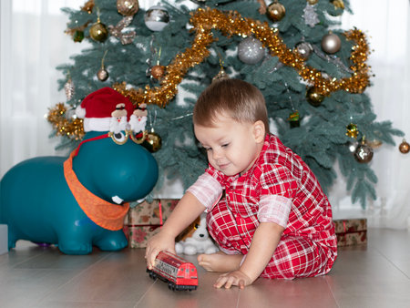 Christmas concept. New Year 2024. A small handsome boy in red checkered pajamas sits on the floor and plays with a red steam locomotive against the background of a decorated Christmas tree with gifts.の写真素材