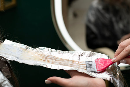 Beauty sphere. Hair coloring in a beauty salon. A master hairdresser-colorist dyes a clients brown hair blond. Apply lightening powder to hair onto foil using a pink brush. Close-up.の写真素材