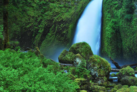 Blue waterfall flowing down into a green valley filled with rocks and plantsの写真素材