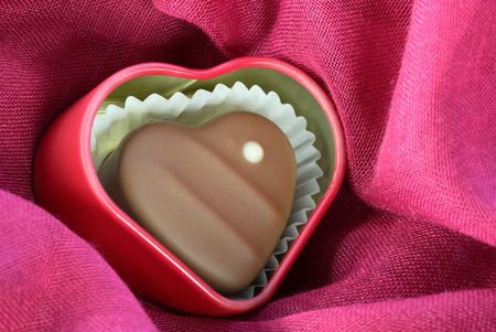 Chocolate-covered heart with a white dot on it, inside a red heart-shaped tin, on pink fabric. Horizontal shot.の写真素材