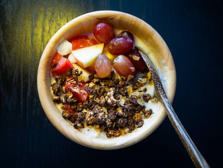 The top shot on the wooden cup of yogurt with fruite and oat meal and wooden spoon that placed on the black table. It is an easy meal for social distancing and COVID lockdownの写真素材