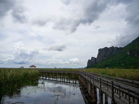 The wooden bridge that connect to the center of the lotus pond and cloudy sky and tall mountain in Thailandの写真素材
