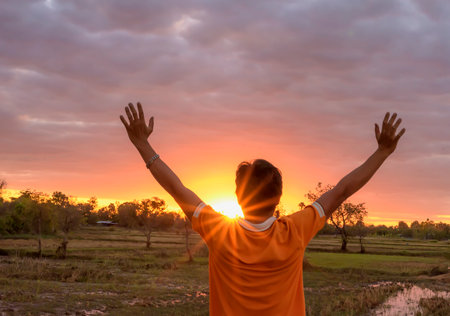 Silhouette of a man raising his hand to watch the sunset or sunrise on a summer field against a cloudy sky background.の写真素材