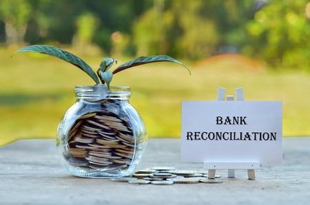 Business Concept - Money in glass container with tree and white board written Bank Reconciliationの写真素材