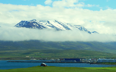The distance view of a small house overlooking the snowy and foggy mountain and blue ocean in the summer along the Ring Road in Icelandの写真素材
