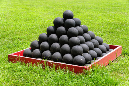 A pyramid of cannonballs, resting in a red wooden crate, sits amidst a vibrant green lawn near Fort...