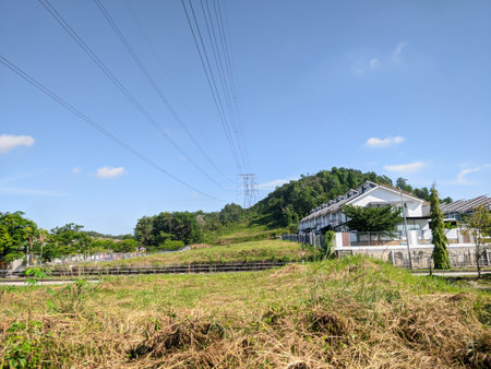 A vibrant landscape under a clear blue sky, featuring power lines and a transmission tower above lush green hills. Modern terraced houses blend into the natural environment, bathed in bright sunlight.の写真素材