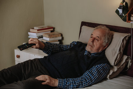 Senior man napping while lying down in bed and watching TV with a television remote, resting in a bedの写真素材