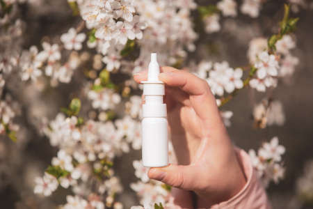 Allergy concept, young woman with nose or nasal spray in hand in front of blooming a tree during spring season, healthcareの写真素材