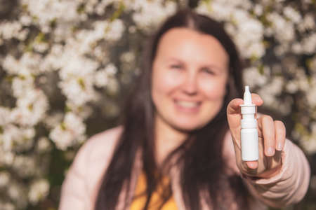 Allergy concept, young woman with nose or nasal spray in hand in front of blooming a tree during spring season, healthcareの写真素材
