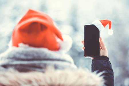 Young woman taking photo of snowy nature by a smartphone with santa hatの写真素材