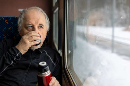 Senior man enjoying fresh tea during travel by train during winter time, traveling conceptの写真素材