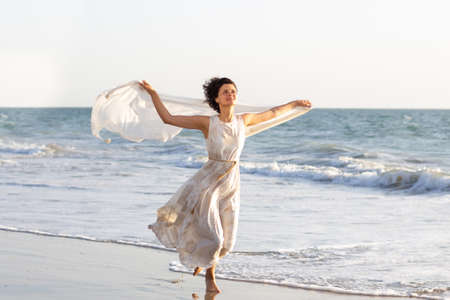 a beautiful girl in a white dress walks along the seashore with a pareo in her handsの写真素材