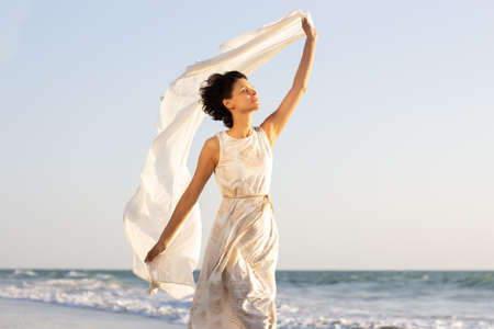 a beautiful girl in a white dress walks along the seashore with a pareo in her handsの写真素材
