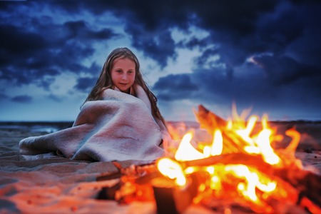 A young girl on the beach at night wrapped in a blanket is heated by the fire.の写真素材