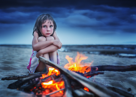 Desperate child hugging her knees with tears in his eyes, night sitting on the beach campfire.の写真素材