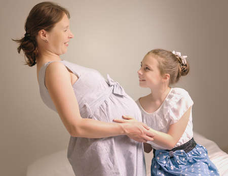 Beautiful pregnant woman and her cute little daughter are smiling while spending time together at home.の写真素材