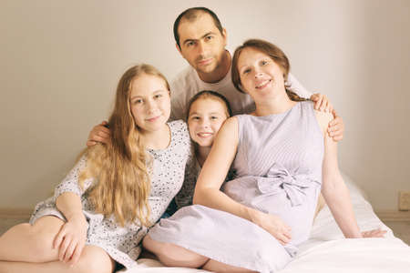 Cute girls and her handsome dad are hugging her beautiful pregnant moms tummy, looking at camera and...