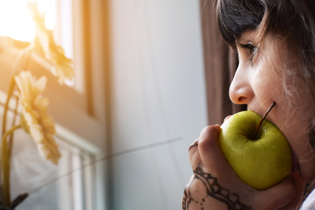 Closeup of Beautiful little Girl child kid portrait eating green healthy Apple while looking outside from home window on sunny day in eveningの写真素材