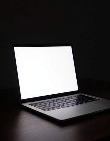 Laptop with blank screen on wooden table in dark room, stock photo