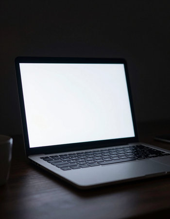Laptop with blank screen on desk in dark room, stock photo