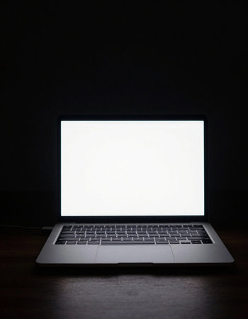Laptop with blank screen on wooden table in dark room, stock photo