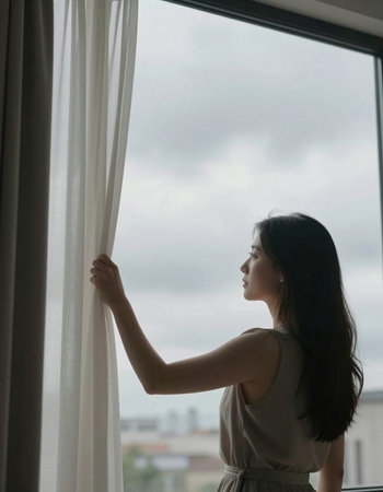 Asian woman looking out the window in the morning with cloudy sky background