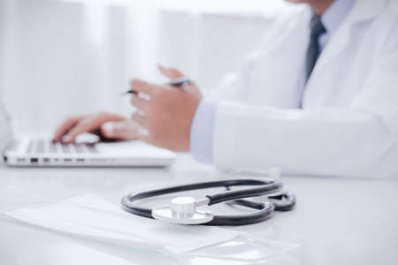 Doctor working with laptop computer and writing on paperwork. Hospital background. Healthcare and medical concept. Focus on stethoscopeの写真素材