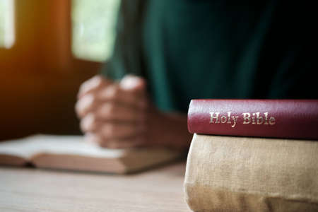 Man with Bible praying, hands clasped together on her Bible on wooden table.