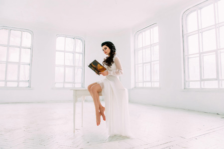 Luxurious brunette in a white dress standing in a white clean room with light and holding a book , a fashionable toning , creative colorの写真素材